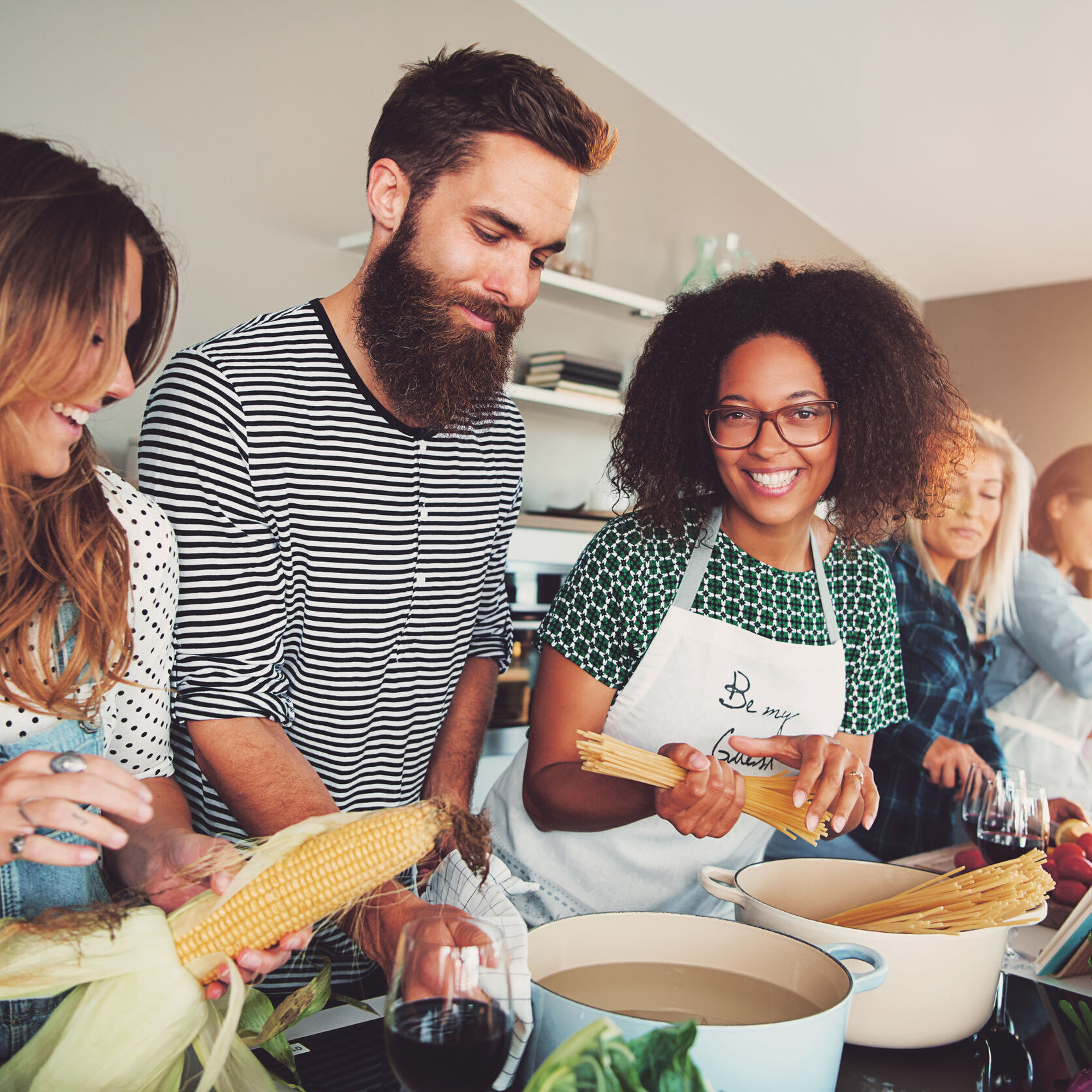 Happy group of young adult men and women cooking together at home in small kitchen or culinary classroom